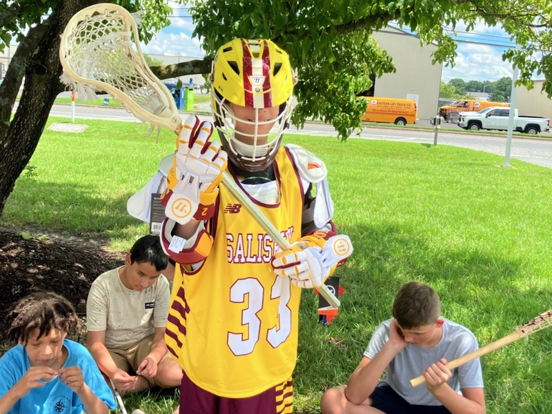 Aaron stands in shade of tree wearing lacrosse helmet, pads, and gloves with stick held in two hands with other campers sitting.