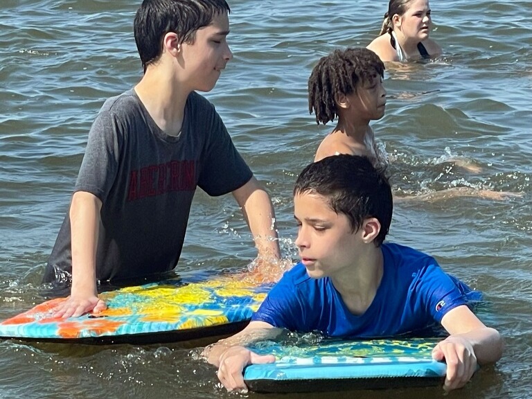 Aaron and Adam on boogie boards in lake with other campers in background.