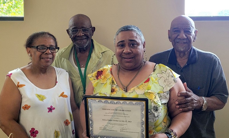 Debra, in a floral print dress, is surrounded by her family as she displays her ITR graduation certificate.
