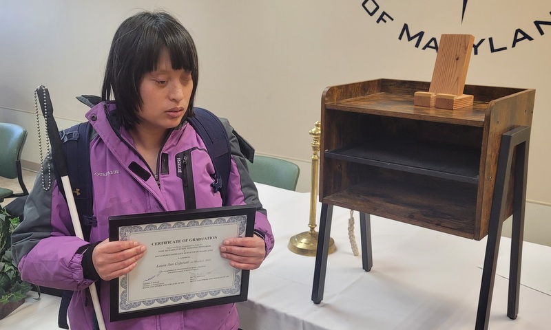 Sunny – posing for the camera with her CORE certificate of completion. Sunny is wearing a purple jacket, holding her cane and standing next to the iPad stand she made in woodshop.