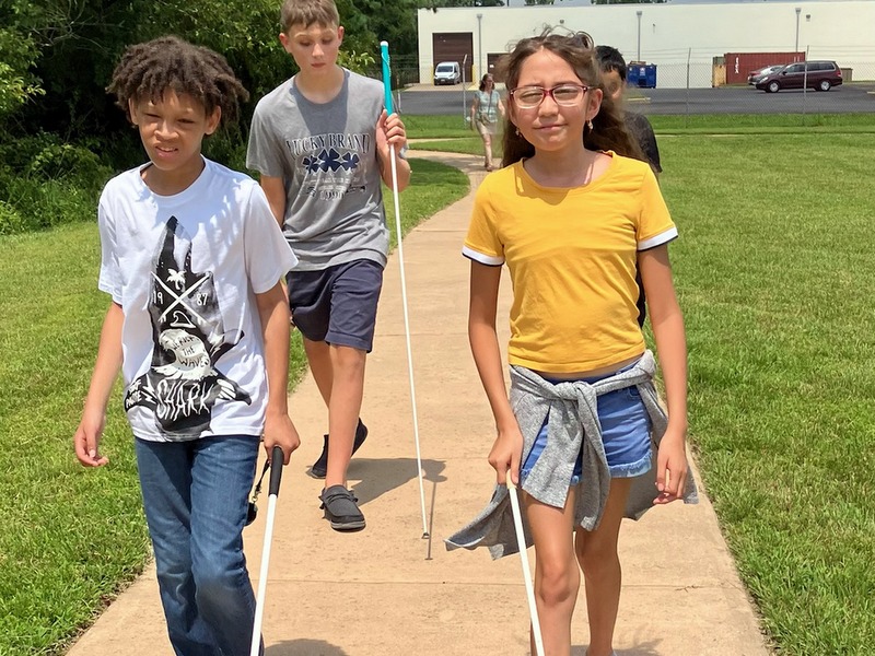 Group of three campers practice cane travel along sidewalk. Wearing short-sleeve shirts, the youth enjoy a sunny day.