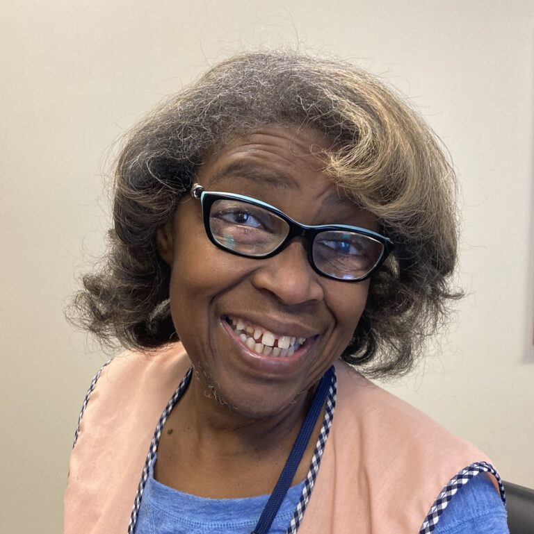 Photo showing Deborah smiling happily at the camera. She has curly dark hair and wears a blue shirt covered by a beige work apron.