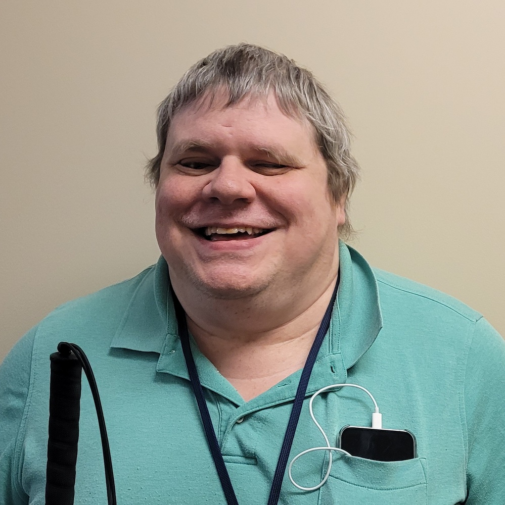 Kyle Seitz, wearing a green shirt with a cellphone in the pocket and holding a cane, smiles joyfully into the camera.