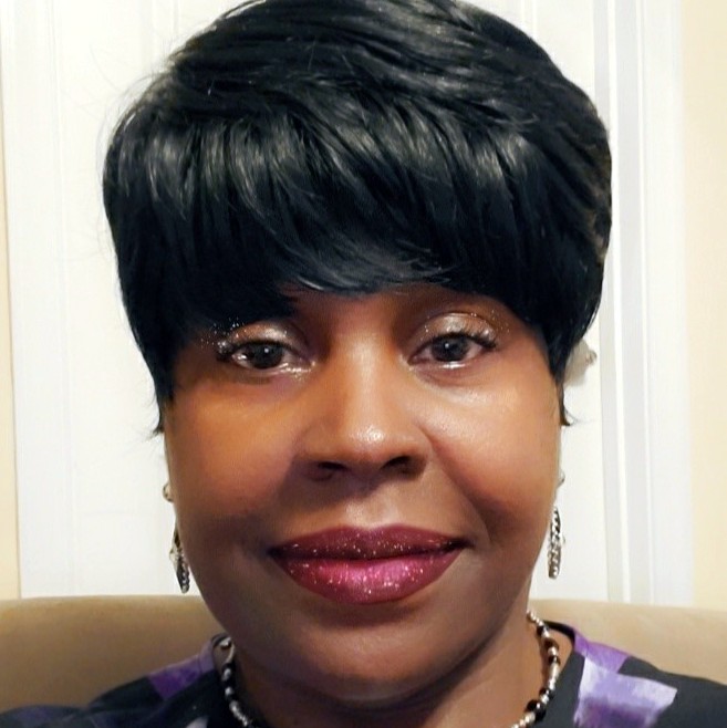 Photo shows a smiling Sheila from the chest up. She sports a short haircut and bold red lips. Earrings dangle from her ears and she wears a long necklace with a gold and black pendant at the end to match her purple, gray, and black top.