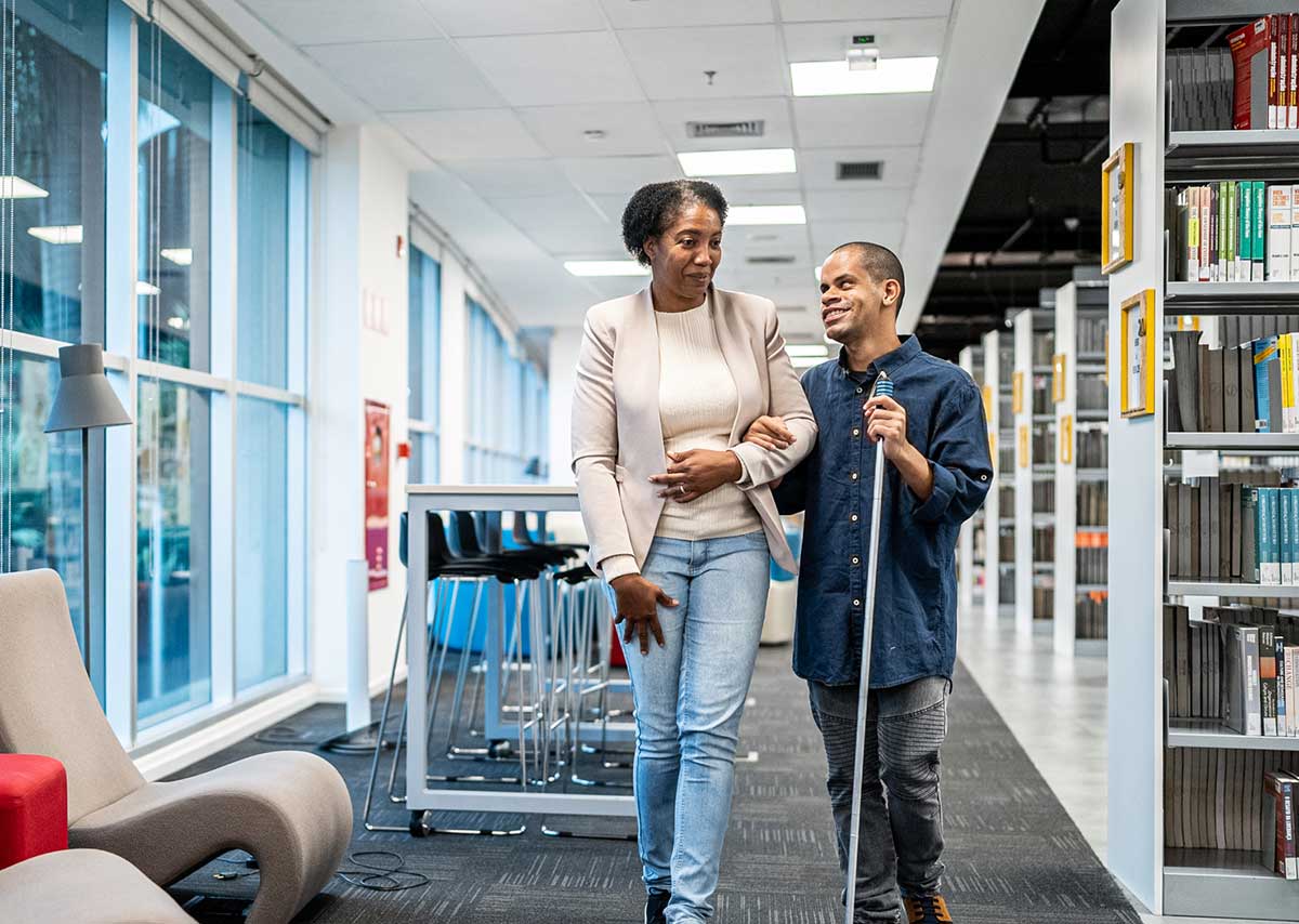 Two adults enjoying conversation while navigating through library. Man is using cane and holding woman's arm.