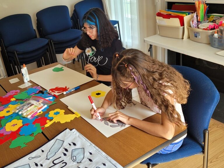 Alexa and Alixon create poster from the many art supplies on the tables in front and behind them.