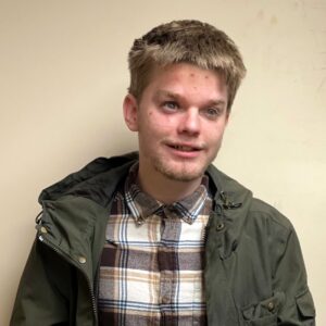 James, wearing a plaid shirt and dark olive jacket, smiles into the camera.