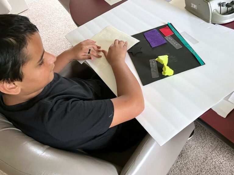 Young camper relaxes in chair at table full of craft supplies. The boy, wearing a black shirt, is creating a poster.