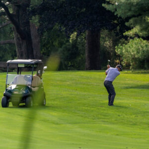 Male golfer taking a shot, standing next to a golf cart on a lush green fairway.