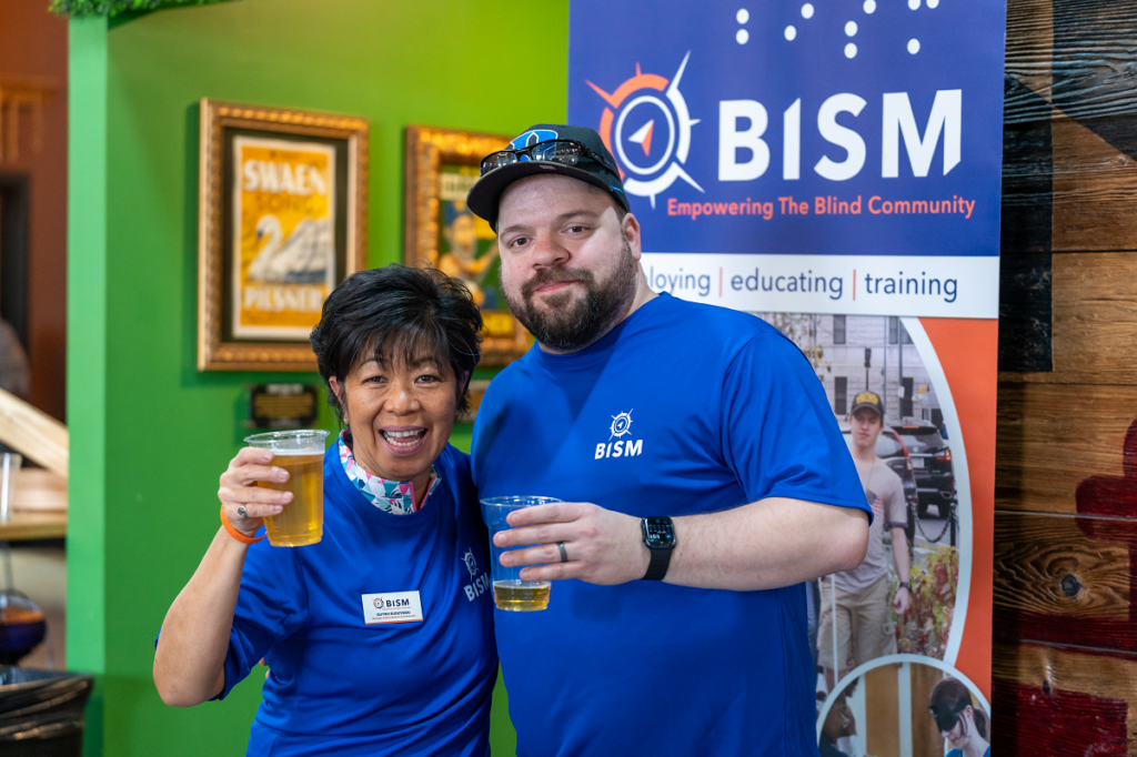 Two BISM team members smile and raise their beers while wearing blue BISM shirts at the BISM on Tap event. A BISM banner stands behind them with the words "Empowering the Blind Community."