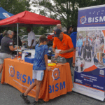 A BISM employee demonstrates a braille typewriter to a young boy at the BISM booth during Frederick Road Fridays. The booth features an orange BISM tablecloth, informational materials, and a large banner showcasing BISM’s mission of empowering the blind community. Other volunteers and community members are visible in the background under red and blue tents.
