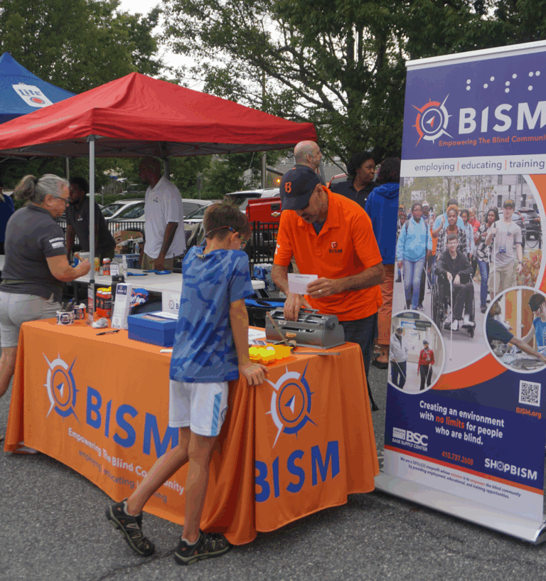 A BISM employee demonstrates a braille typewriter to a young boy at the BISM booth during Frederick Road Fridays. The booth features an orange BISM tablecloth, informational materials, and a large banner showcasing BISM’s mission of empowering the blind community. Other volunteers and community members are visible in the background under red and blue tents.