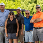 Group photo of five people standing outdoors at the BISM Pro-AM Golf Classic. From left to right: the golf pro, Kimberly Kennedy from SP Richards, Sean from SP Richards, Jeremy Fisher from SP Richards, and Mike from BISM. All are smiling and wearing golf attire, standing in front of a golf cart with trees in the background.