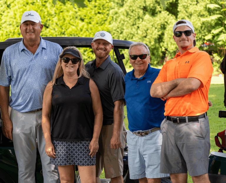 Group photo of five people standing outdoors at the BISM Pro-AM Golf Classic. From left to right: the golf pro, Kimberly Kennedy from SP Richards, Sean from SP Richards, Jeremy Fisher from SP Richards, and Mike from BISM. All are smiling and wearing golf attire, standing in front of a golf cart with trees in the background.
