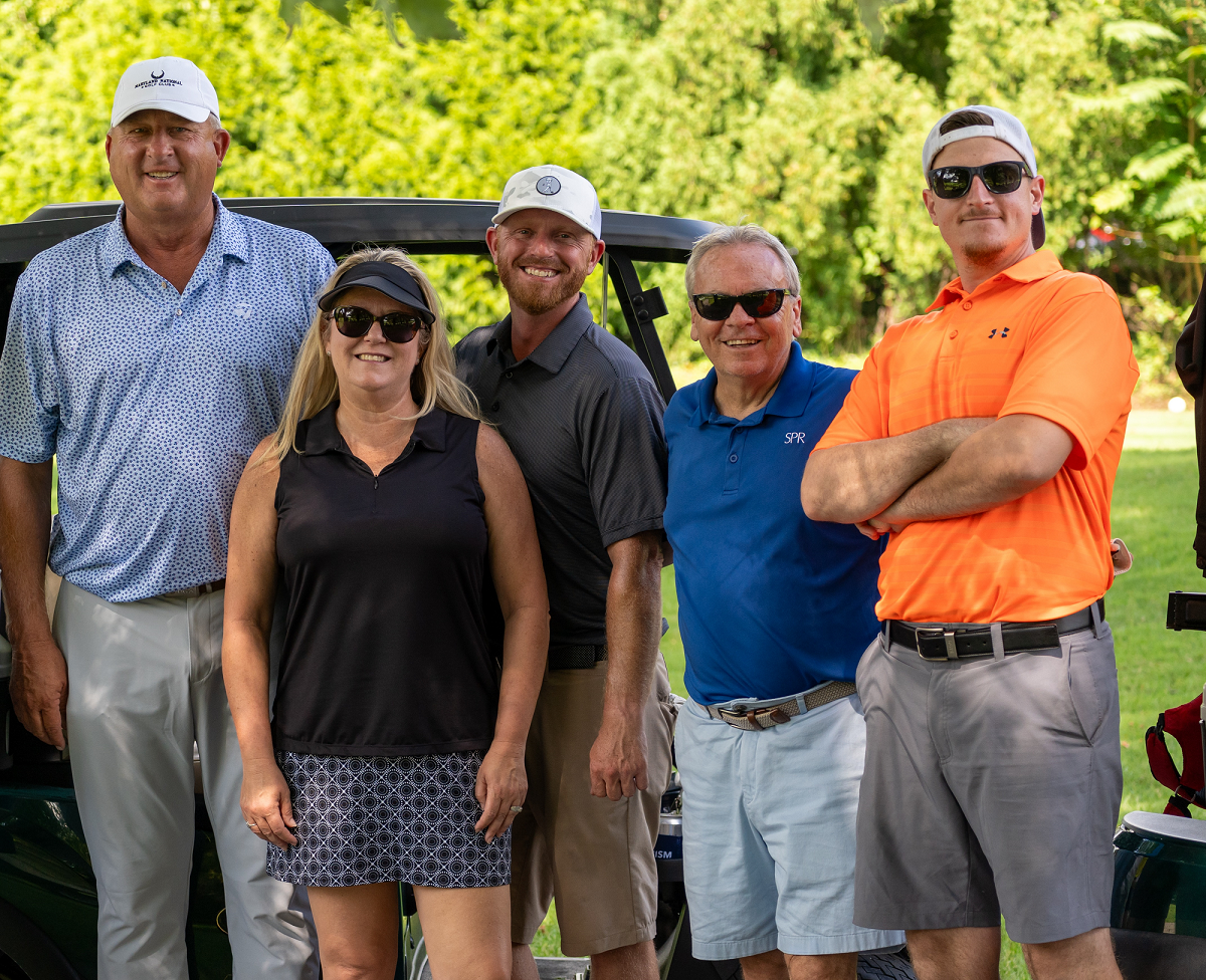 Group photo of five people standing outdoors at the BISM Pro-AM Golf Classic. From left to right: the golf pro, Kimberly Kennedy from SP Richards, Sean from SP Richards, Jeremy Fisher from SP Richards, and Mike from BISM. All are smiling and wearing golf attire, standing in front of a golf cart with trees in the background.