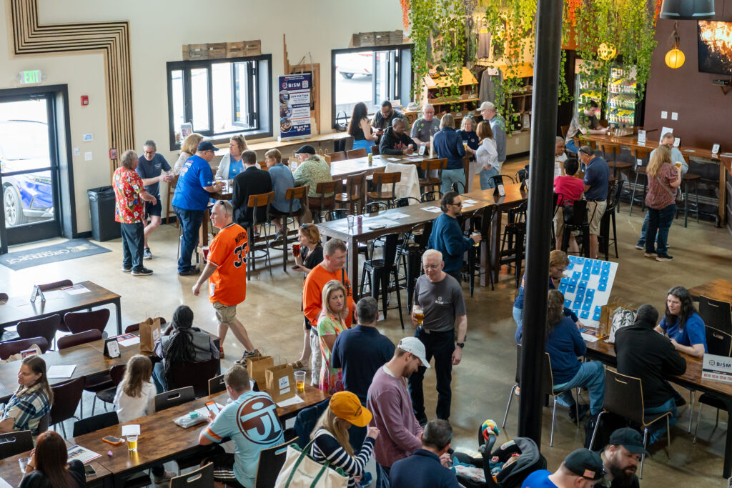 A vibrant scene inside Checkerspot Brewing Company during BISM’s community event. The brewery is filled with attendees of all ages mingling, enjoying drinks, and participating in activities. Tables are occupied by guests chatting and sampling food and drinks, while others explore games and displays, including a BISM-themed game board. Greenery hangs from the ceiling, creating a welcoming atmosphere. The space is lively and full of energy, showcasing strong community engagement and support.