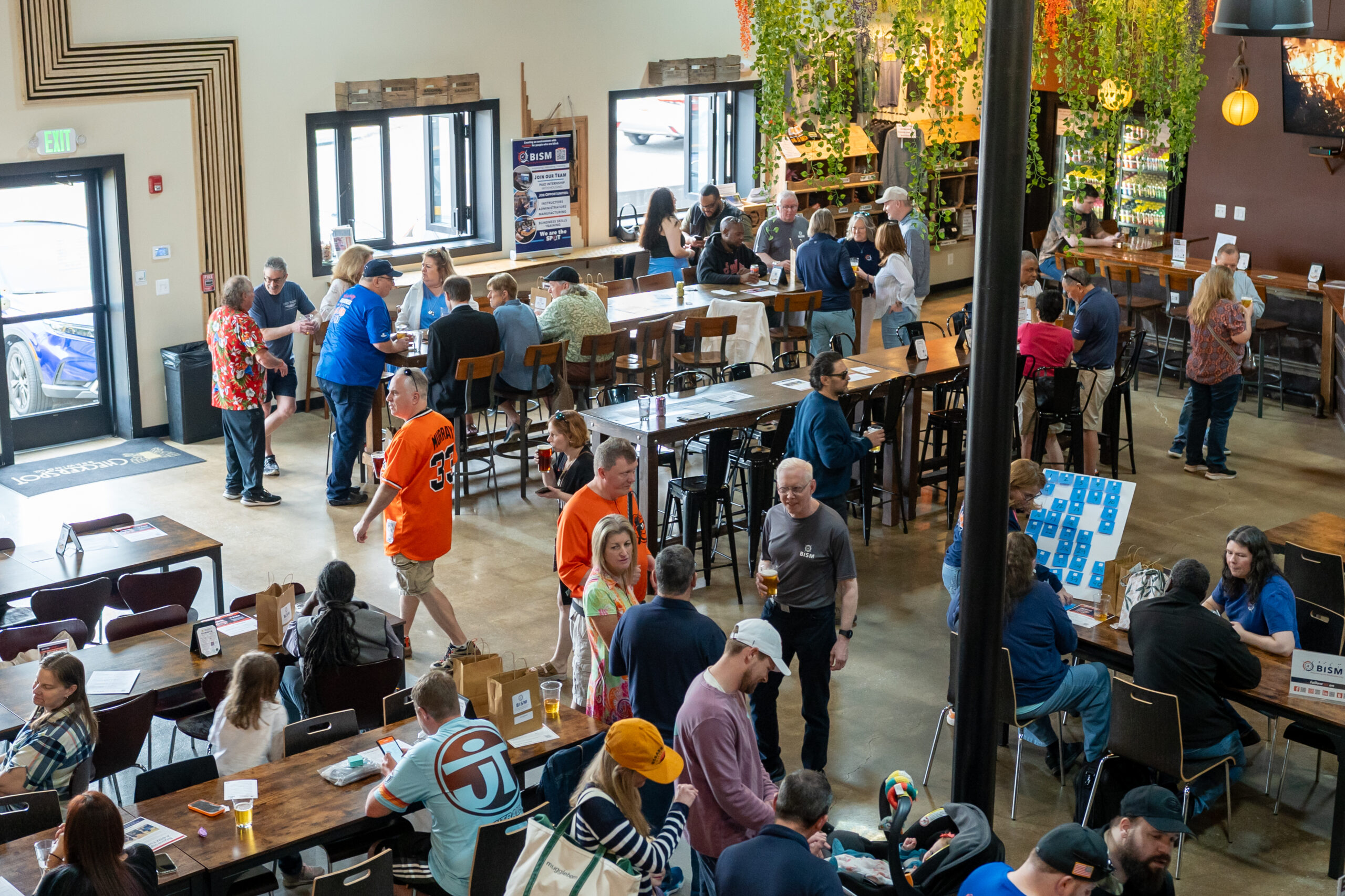 A vibrant scene inside Checkerspot Brewing Company during BISM’s community event. The brewery is filled with attendees of all ages mingling, enjoying drinks, and participating in activities. Tables are occupied by guests chatting and sampling food and drinks, while others explore games and displays, including a BISM-themed game board. Greenery hangs from the ceiling, creating a welcoming atmosphere. The space is lively and full of energy, showcasing strong community engagement and support.