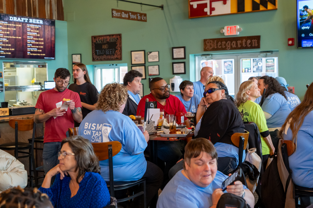 A lively crowd gathers inside a brewery during a BISM on Tap event. Attendees sit and stand around tables enjoying food, drinks, and conversation. Several people wear matching light blue BISM on Tap staff shirts, while others socialize throughout the space. The brewery features wooden décor, beer menus, and signs hanging on the walls, creating a warm and energetic community atmosphere.