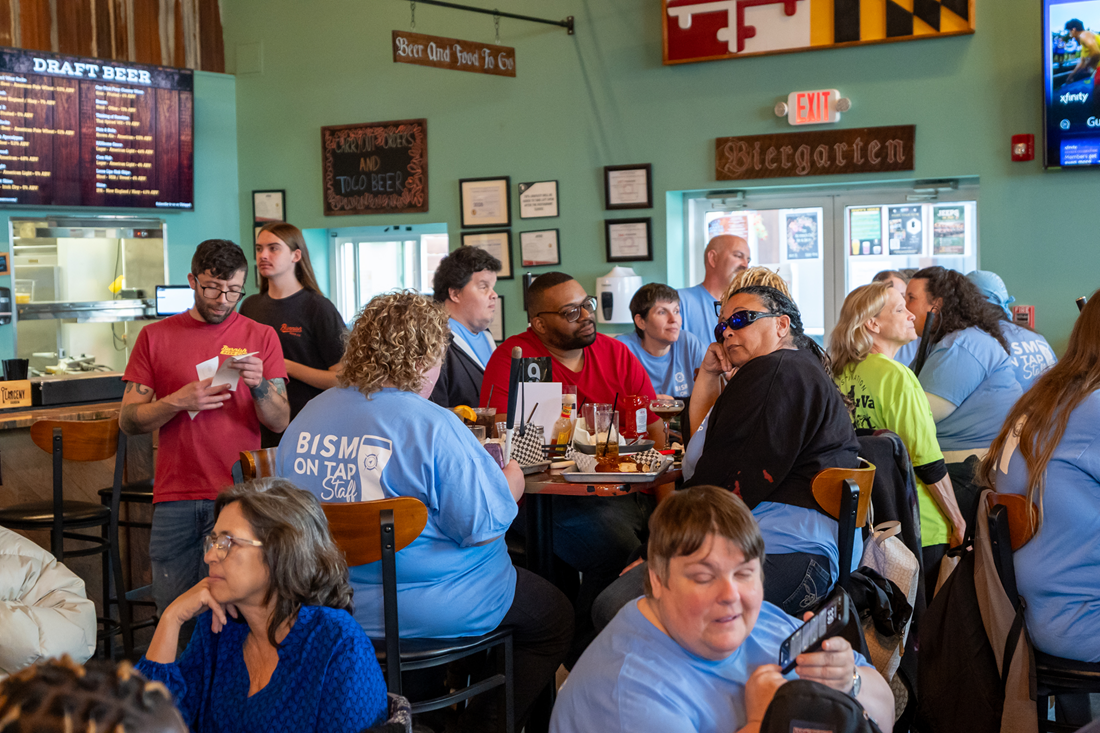 A lively crowd gathers inside a brewery during a BISM on Tap event. Attendees sit and stand around tables enjoying food, drinks, and conversation. Several people wear matching light blue BISM on Tap staff shirts, while others socialize throughout the space. The brewery features wooden décor, beer menus, and signs hanging on the walls, creating a warm and energetic community atmosphere.
