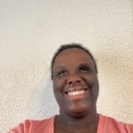 A smiling Black woman in a coral shirt stands in front of a neutral wall.
