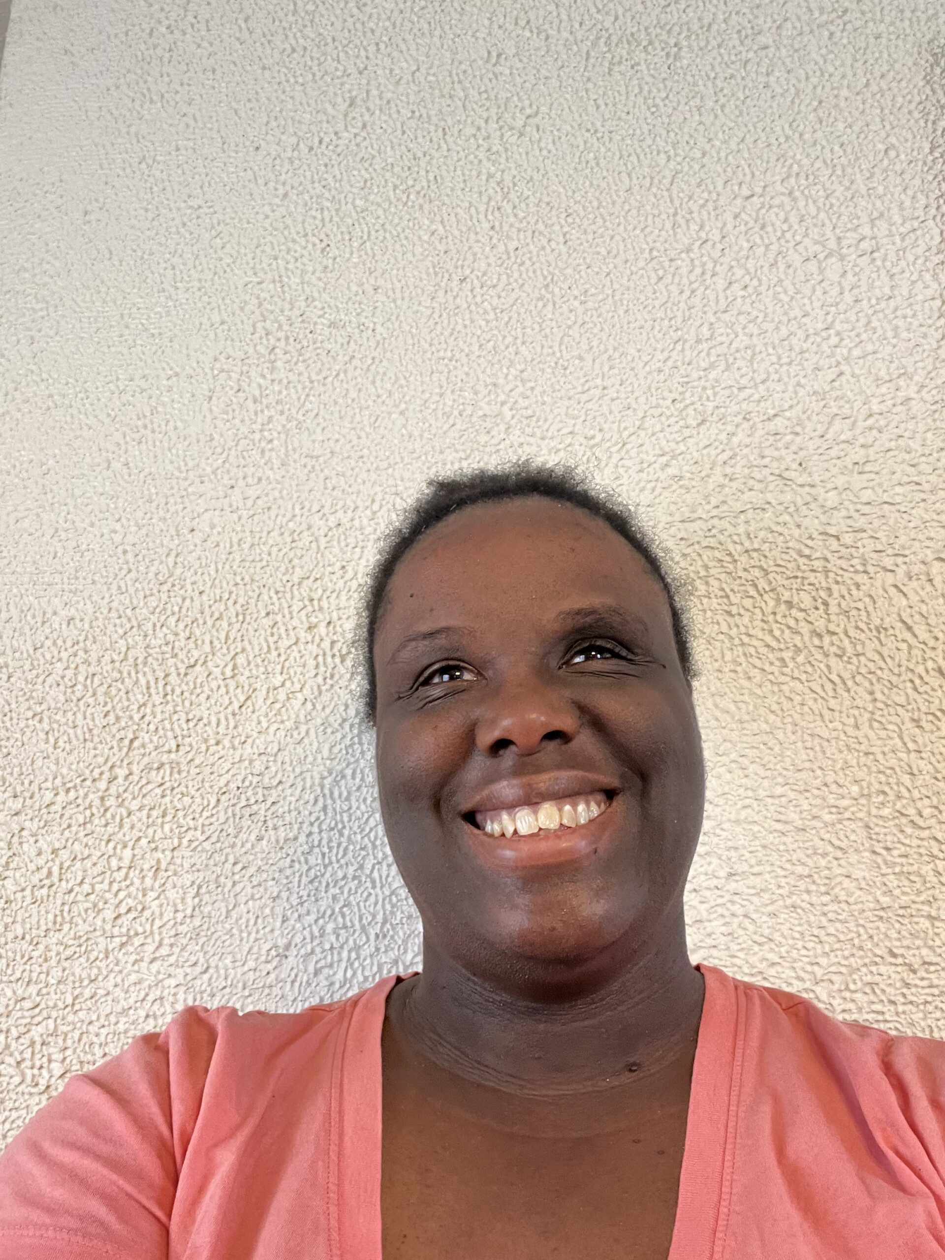 A smiling Black woman in a coral shirt stands in front of a neutral wall.