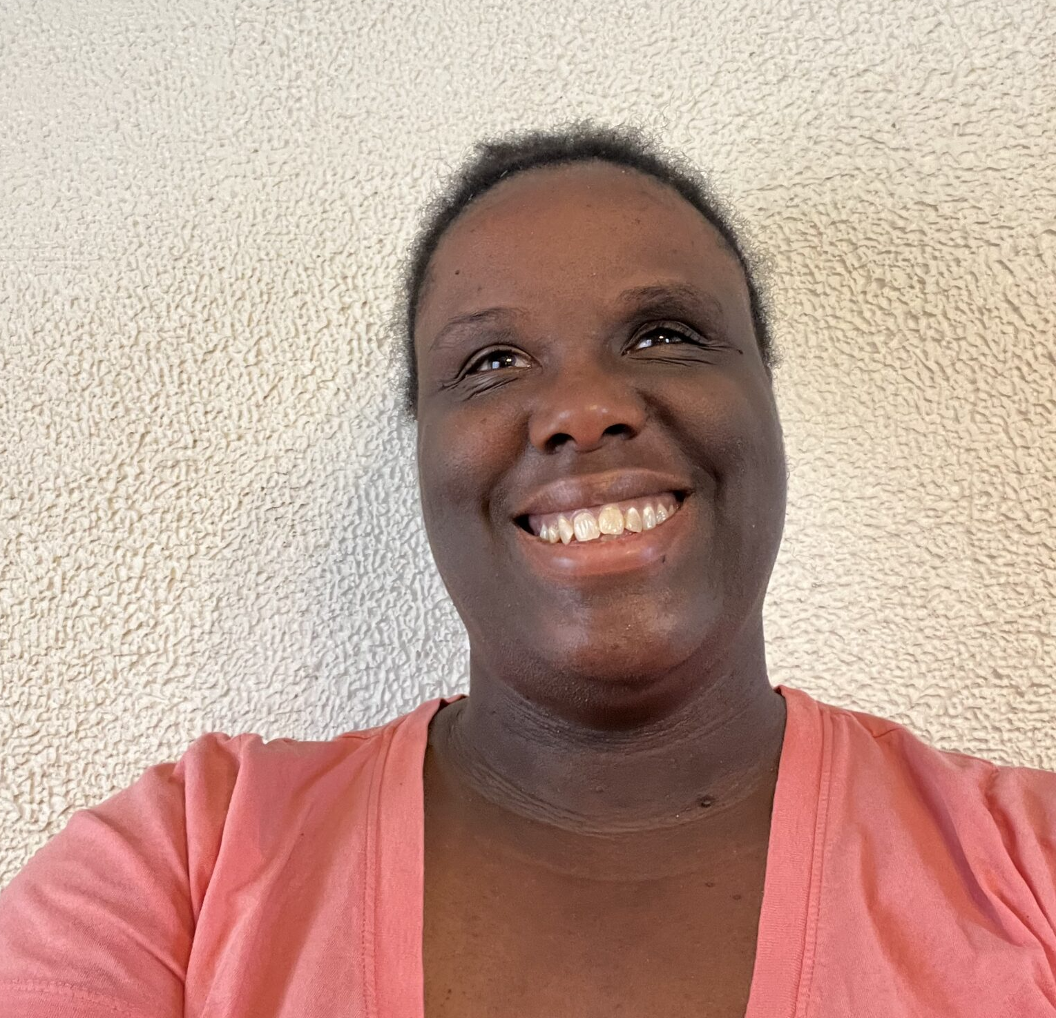 A smiling Black woman in a coral shirt stands in front of a neutral wall.