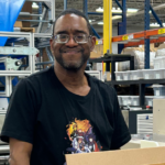 A smiling Black man wearing glasses and a black t-shirt sits at a workstation with boxes and equipment in a warehouse setting.