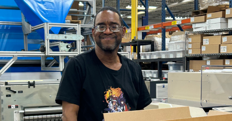 A smiling Black man wearing glasses and a black t-shirt sits at a workstation with boxes and equipment in a warehouse setting.