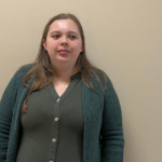 A white woman with long brown hair stands against a plain beige wall, wearing a green cardigan over a matching button-up shirt, looking toward the camera with a neutral expression.
