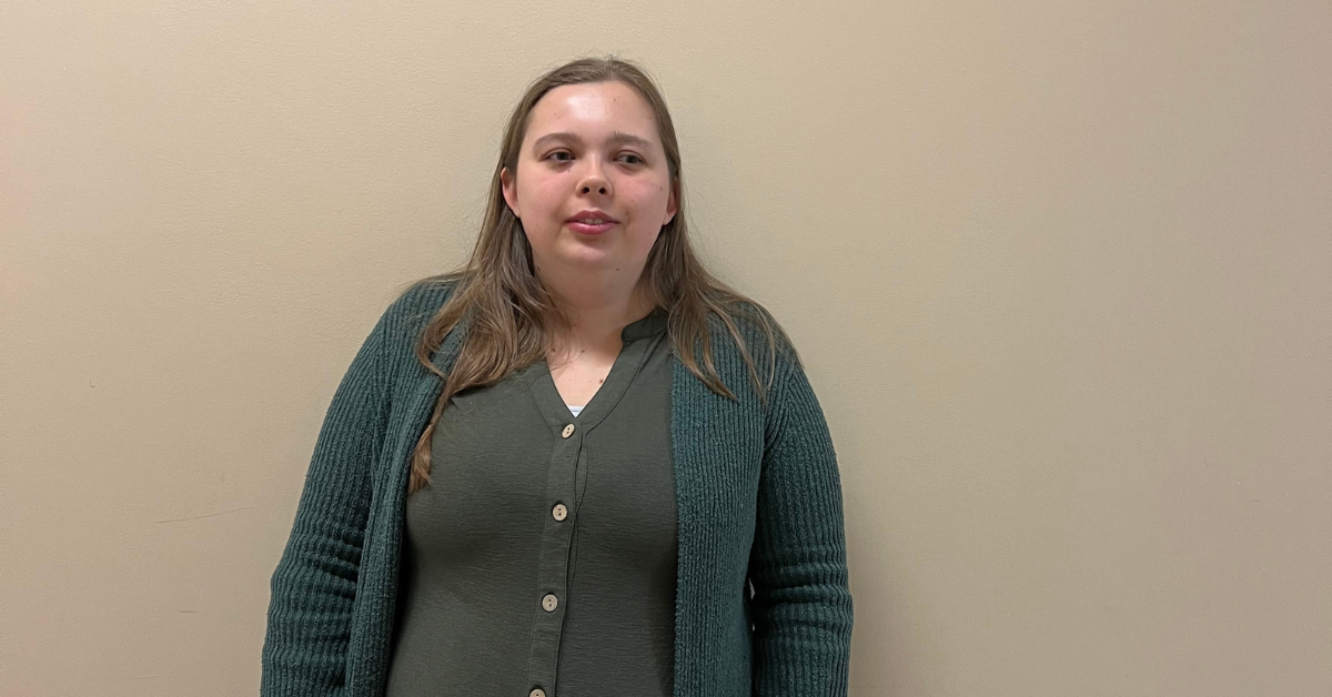 A white woman with long brown hair stands against a plain beige wall, wearing a green cardigan over a matching button-up shirt, looking toward the camera with a neutral expression.