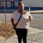 A Black woman standing on a city sidewalk holding a white mobility cane.