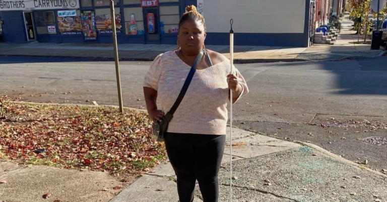 A Black woman standing on a city sidewalk holding a white mobility cane.