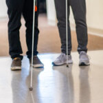 A close up photo of two people standing so that the photo only shows their legs and their white canes.
