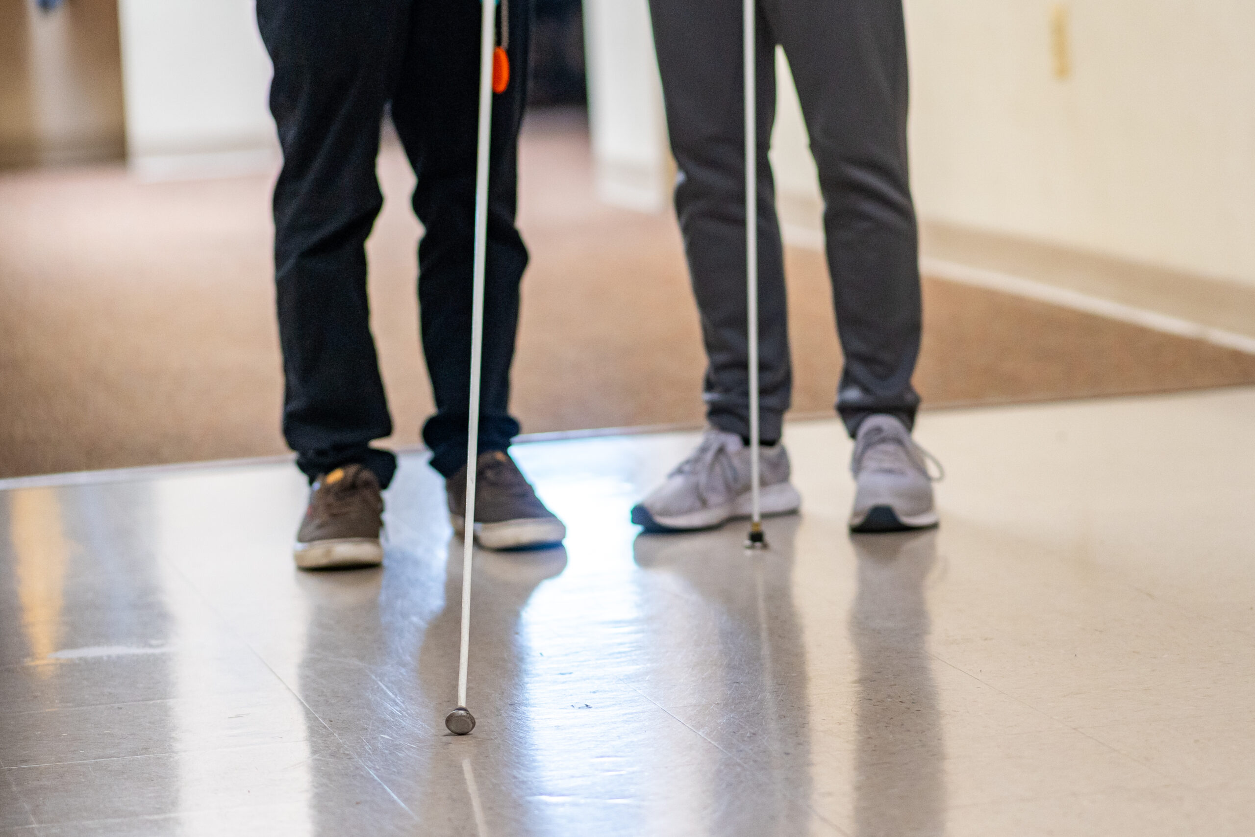A close up photo of two people standing so that the photo only shows their legs and their white canes.